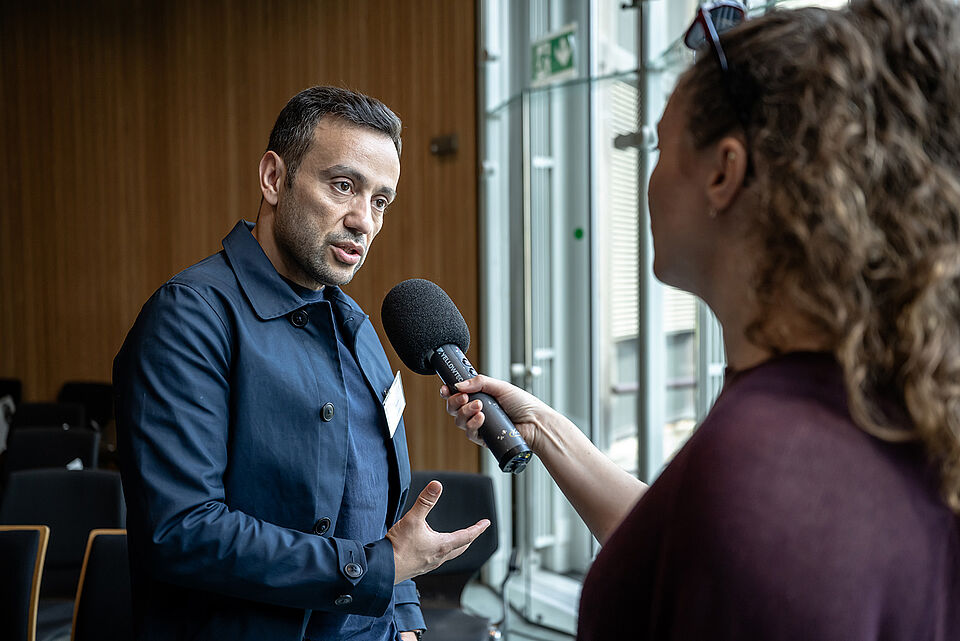 Kouras Interview Ein Mann wird von einer Reporterin mit einem Mikrofon interviewt.