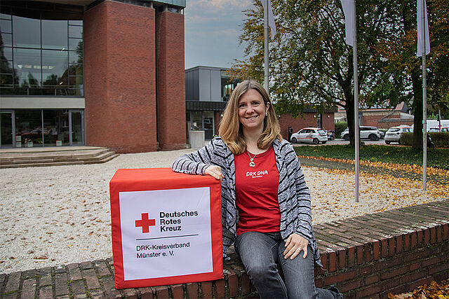 Eine Person sitzt auf einer gemauerten Kante neben einer Box mit der Aufschrift „Deutsches Rotes Kreuz DRK-Kreisverband Münster e. V.“. Die Person trägt ein rotes Shirt mit weißem „DRK DNA“-Schriftzug. Im Hintergrund sind ein modernes Gebäude mit großen Fenstern, Bäume und parkende Autos zu sehen.