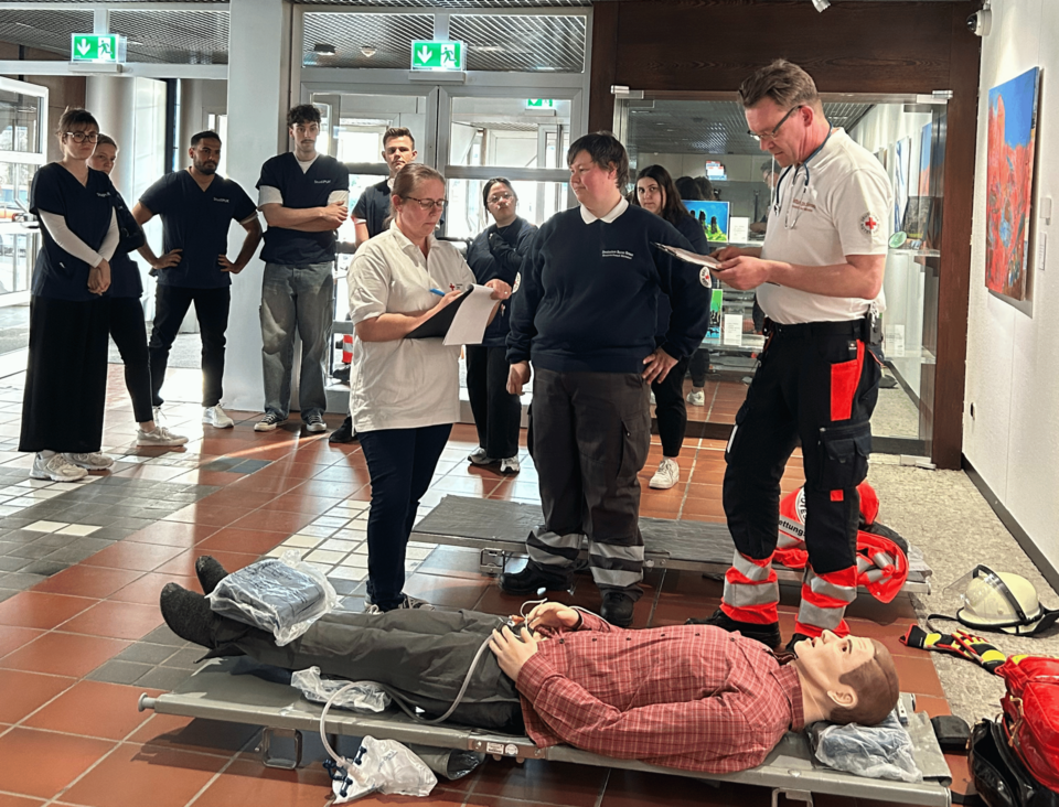 Vorne im Bild liegt eine Puppe auf einer Trage, drei Personen in Einsatzkleidung stehen vor ihr. Im Hintergrund steht eine Gruppe von jungen Personen in blauen Kasacks.