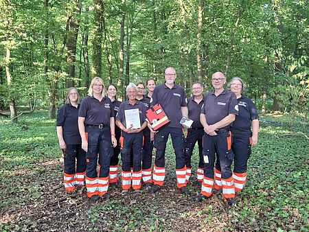 Gruppe von Personen in Einsatzkleidung in einem Wald. Frau in der Mitte hält eine Urkunde in der Hand. Ein Mann eine Mappe und eine Box.