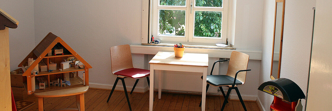 Kinderschutzambulanz Kleiner, heller Raum mit Holzfußboden. In der Mitte steht ein weißer Tisch mit zwei Stühlen (ein roter, ein schwarzer). Auf dem Tisch steht ein gelber Becher mit Buntstiften. Links daneben befindet sich ein Puppenhaus mit Miniaturmöbeln und Spielzeug. Durch das Fenster im Hintergrund fällt Tageslicht, draußen ist Grün zu sehen.
