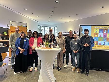 Ein Gruppenbild mit zehn Menschen. Im Hintergrund sind Banner und Leinwände mit dem "Wochen gegen Rassismus" Logo. Im Vordergrund ist ein Stehtisch.