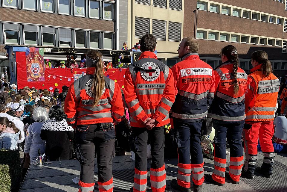 5 Einsatzkräfte mit Einsatzkleidung stehen mit dem Rücken zur Kamera. Sie blicken auf die Straße wo Karnevalswagen lang fahren und sich eine Menschenmenge befindet.