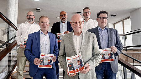 Sechs Männer in Anzügen auf einer Treppe halten eine Broschüre in die Kamera