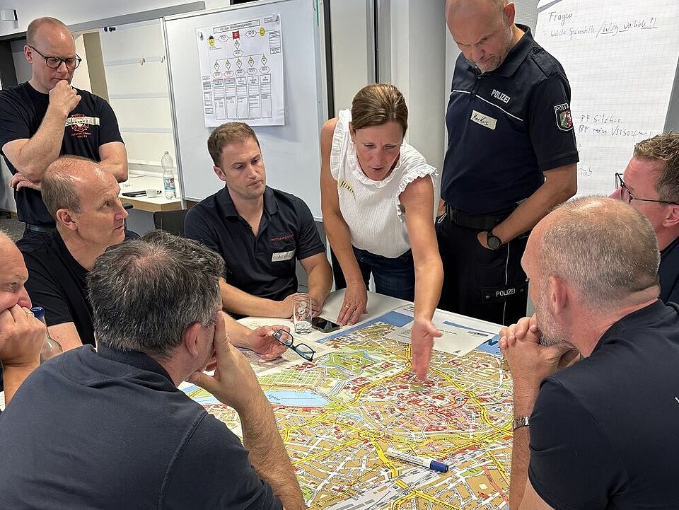 Mehrere Personen, teilweise in Dienstkleidung der Polizei und Feuerwehr, sitzen und stehen um einen Tisch herum, auf dem ein Stadtplan liegt.