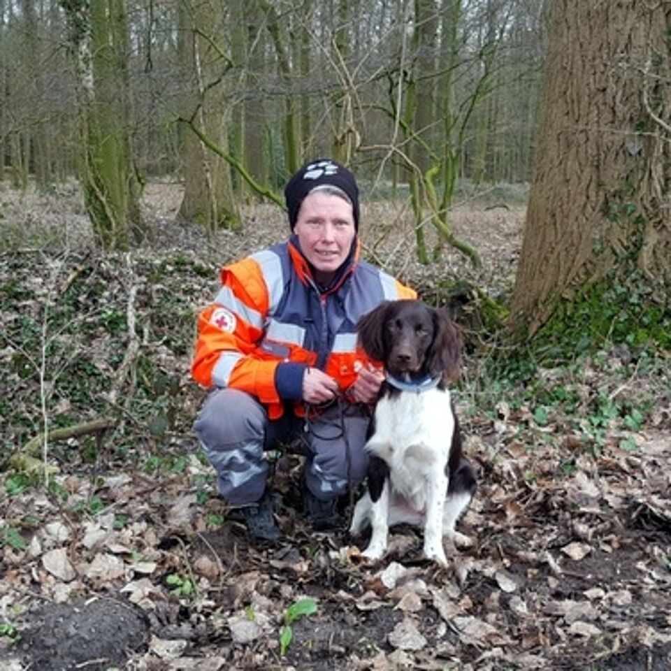 Eine Person in Einsatzkleidung kniet neben einem Hund. Im Hintergrund befindet sich ein Wald.M