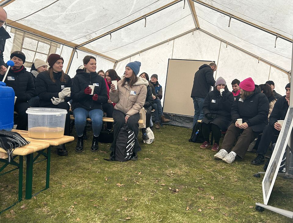 Eine Gruppe von menschen sind auf Bänken in einem Zelt und haben Trinkbecher in der Hand. Vorne steht ein Eimer mit gefiltertem Wasser.