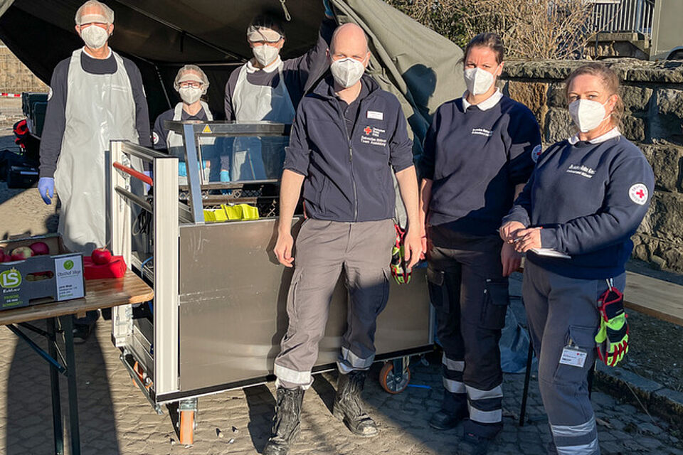 Sechs Personen stehen im Freien neben einem Metallwagen und einem Tisch, auf dem einige Äpfel liegen. Die Personen tragen Einsatzkleidung, einige zusätzlich Schürzen und Handschuhe