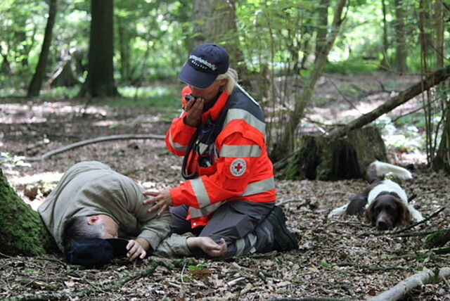 Eine Person in Einsatzkleidung betreut eine andere Person, die am Waldboden liegt. Die betreuende Person nutzt ein Funkgerät zur Kommunikation. In der Nähe liegt ein Hund, der offenbar zur Such- und Rettungsgruppe gehört.