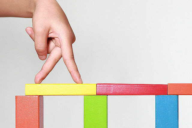Eine aus bunten Holzklötzen gebaute Brücke, über die zwei Finger spazieren.