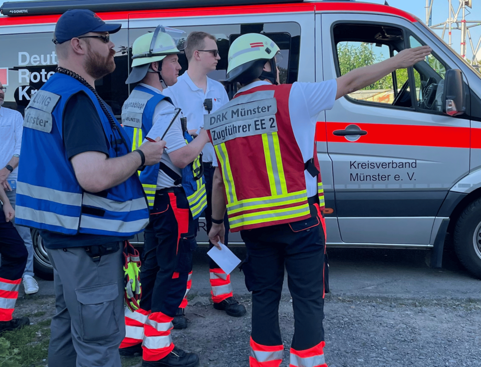 Vier Personen in Einsatzkleidung und Westen mit verschiedenen Zuständigkeiten vor einem Einsatzleitfahrzeug.