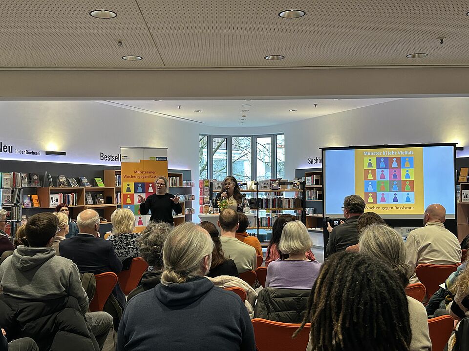 Eine Person steht vor einer Gruppe und hält einen Vortrag. Im Hintergrund ist eine Bibliothek zu sehen und das Logo der Wochen gegen Rassismus