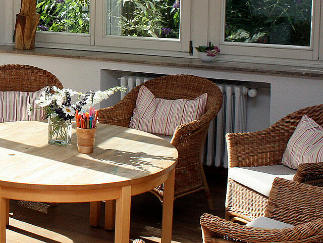 Ein sonnendurchfluteter Raum mit einem runden Holztisch, umgeben von sechs Korbstühlen mit Sitzkissen. Große Fenster auf drei Seiten lassen viel Tageslicht herein. Auf dem Tisch und den Fensterbänken stehen Blumenvasen und Holzskulpturen. Der Boden ist aus Holz, an einer Wand befindet sich ein Heizkörper.
