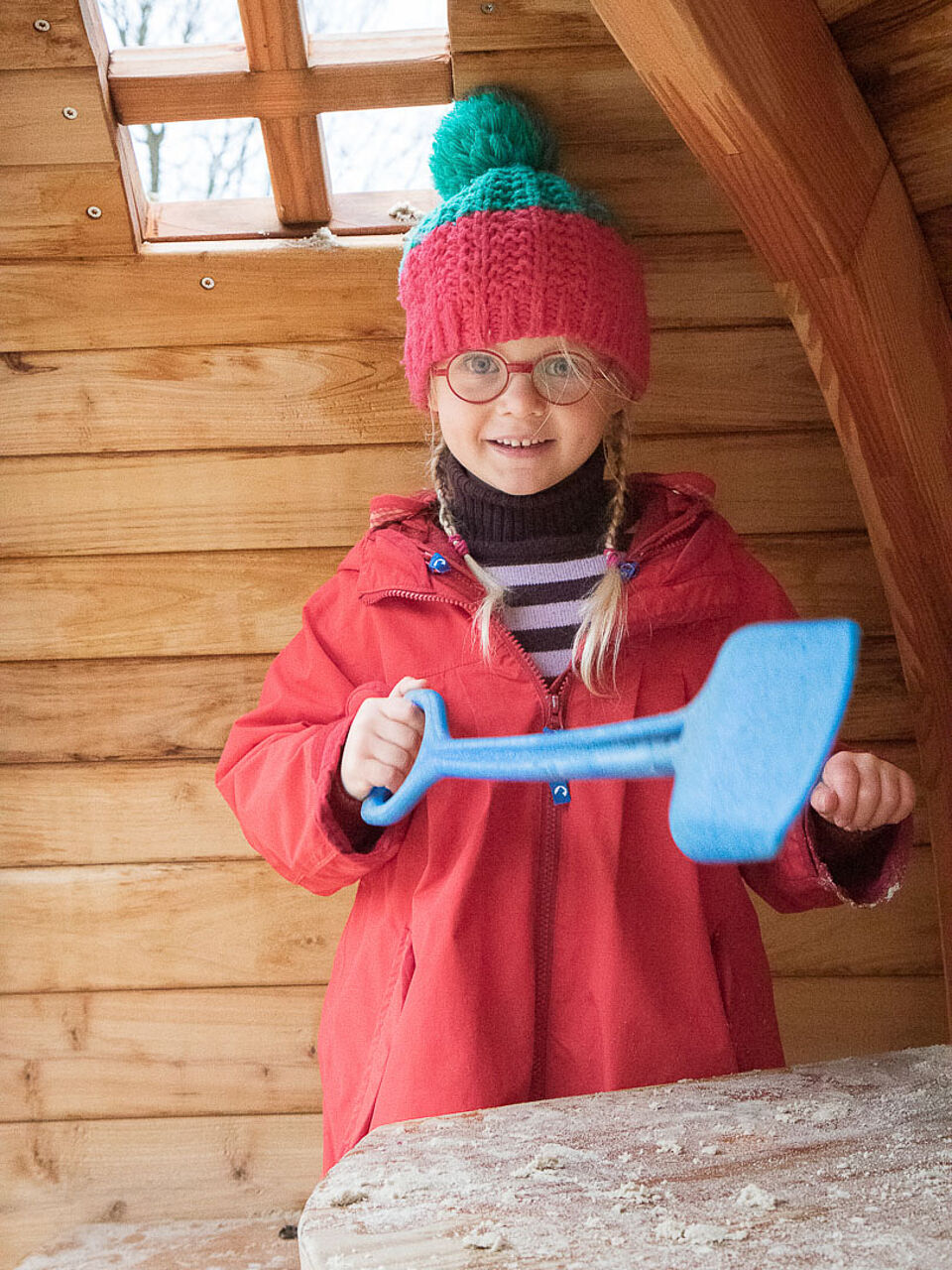 Ein Kinder in warmer Kleidung in einer Holzhütte mit einer blauen Plastikschaufel in der Hand