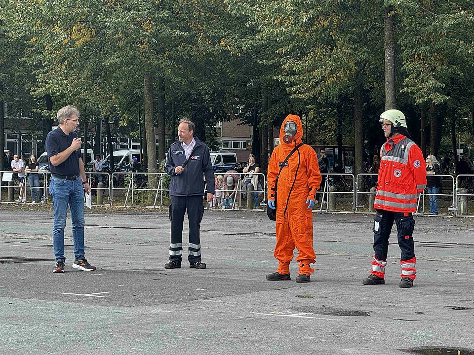 Vier Personen stehen in einer Reihe. Eine Person in Katastrophenschutz-Einsatzkleidung, eine Person in Schutzkleidung, zwei Personen mit Mikrofon.