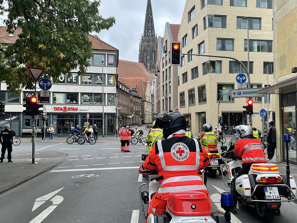 Giro 2025 Einsatzkräfte auf Motorrädern auf einer Kreuzung in der Innenstadt, im Hintergrund Fahrradfahrer*innen und ein Kirchturm