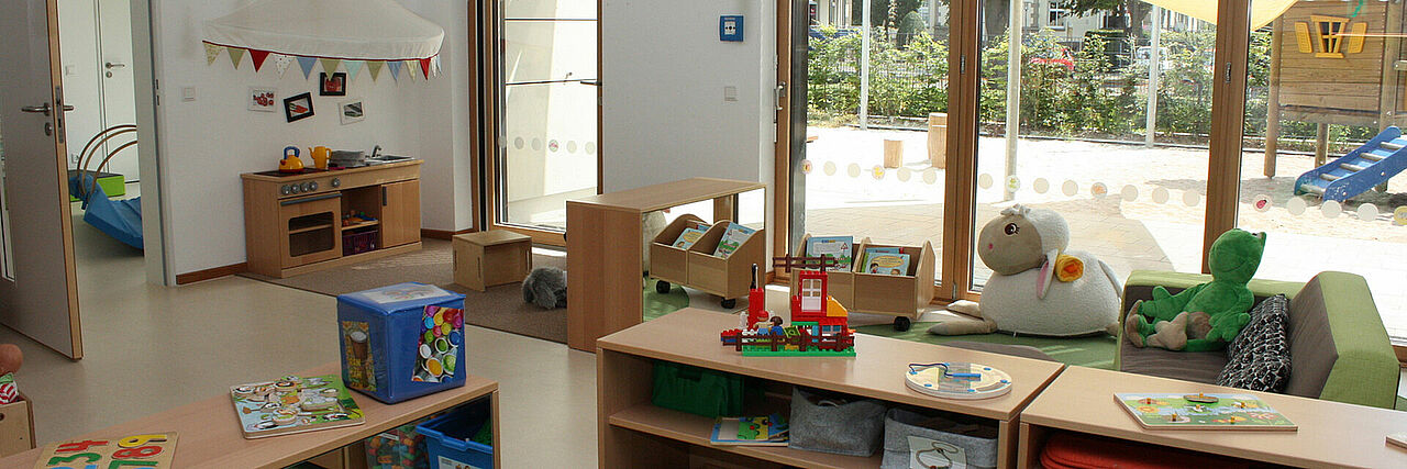 Meerwiese Gruppenraum mit Kindermöbeln und Spielzeug, große Fensterfront wo Außenbereich mit Spielgeräten zu sehen sind