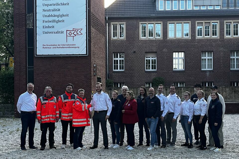 Eine Gruppe von Menschen steht auf einem Kiesplatz. Links stehen drei Personen in Einsatzkleidung, die den anderen Personen eine brennende Fackel überreichen.
