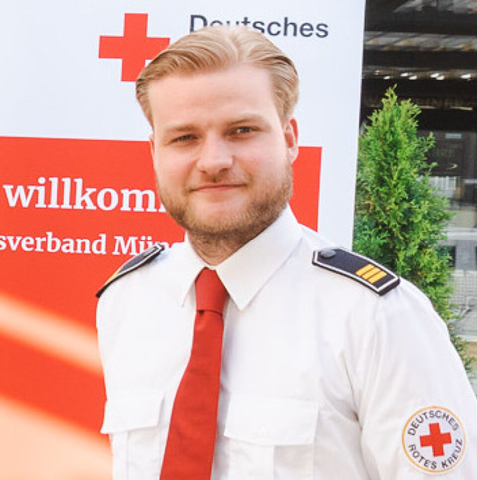 Mann mit blonden Haaren, weißem Hemd mit DRK-Logo und Schulterklappen sowie roter Krawatte, im Hintergrund befindet sich ein Roll-Up vom DRK