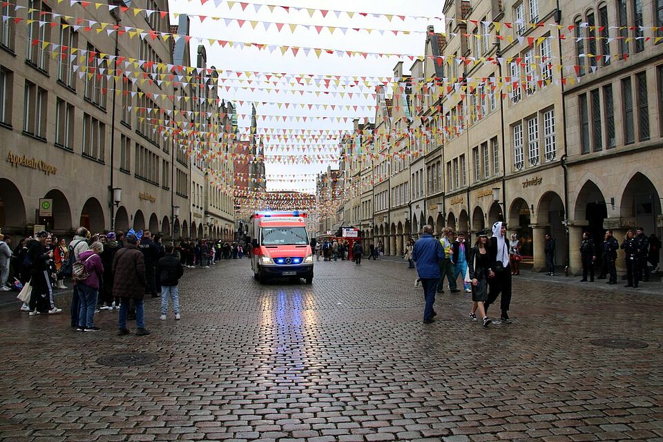 Prinzipalmarkt Straße mit Kopfsteinpflastern, historischen Giebelhäusern und bunten Wimpeln. Zentral auf dem Bild ist ein DRK-RTW mit Blaulicht zu sehen