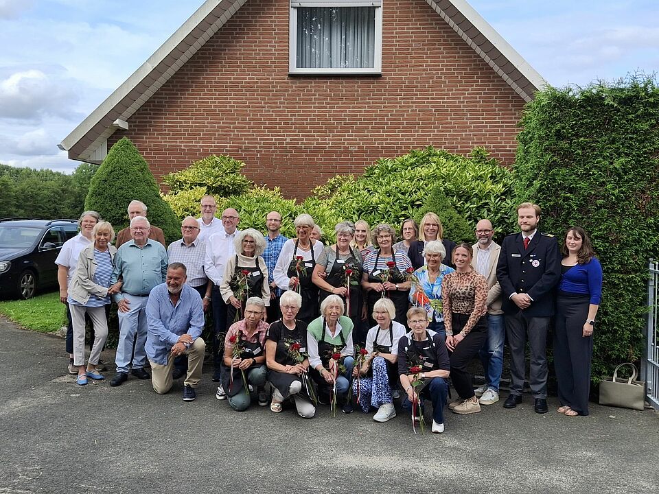 Eine Gruppe von Personen steht vor einer grünen Hecke, im Hintergrund ist ein Haus zu sehen.