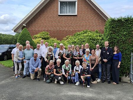 Eine Gruppe von Personen steht vor einer grünen Hecke, im Hintergrund ist ein Haus zu sehen.