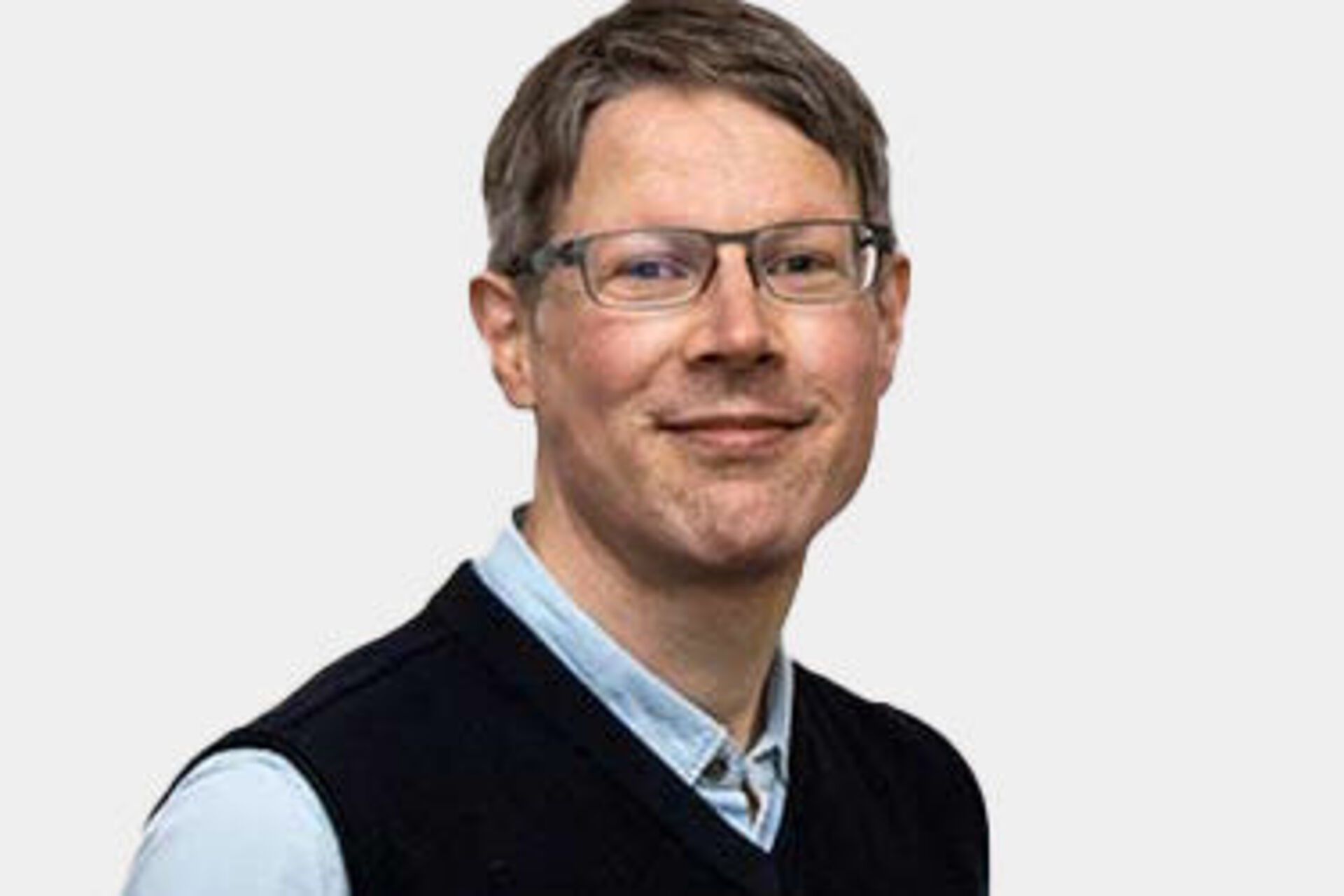 Mann mit Brille und blauem Hemd mit schwarzer Weste vor weißem Hintergrund