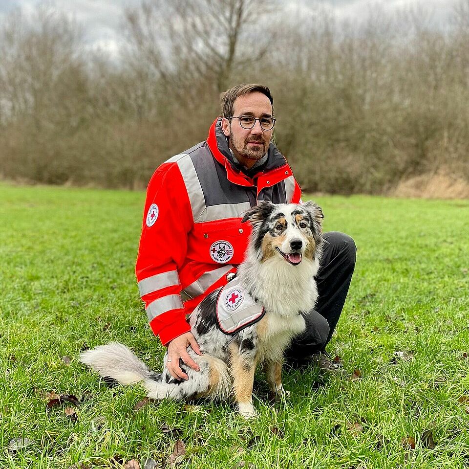 Eine Person in Einsatzkleidung kniet auf einer Wiese. Neben ihr sitzt ein Hund, der eine Weste mit dem Rotkreuz-Symbol trägt. Im Hintergrund sind Bäume und Büsche zu sehen.