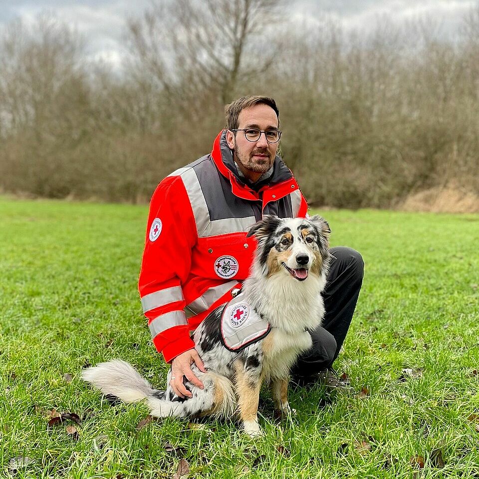 Eine Person in Einsatzkleidung kniet auf einer Wiese. Neben ihr sitzt ein Hund, der eine Weste mit dem Rotkreuz-Symbol trägt. Im Hintergrund sind Bäume und Büsche zu sehen.