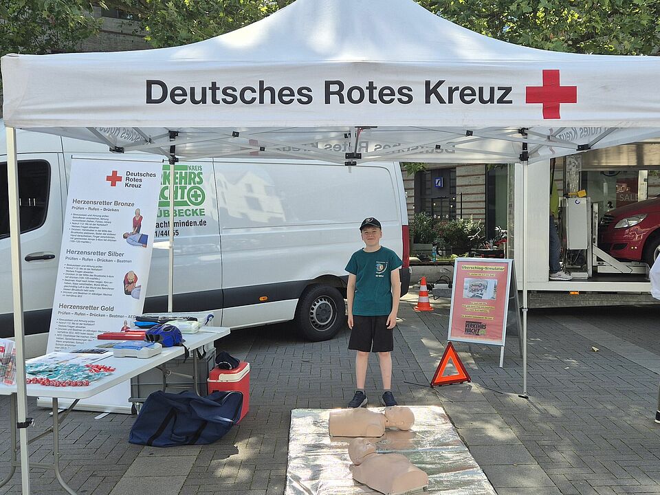 Verkehrssicherheitstag DRK-Pavillion mit Jungen im türkisem T-Shirt, Reanimationspuppen und einem Infostand mit Materialien. Im Hintergrund weitere Stände und Polizisten.