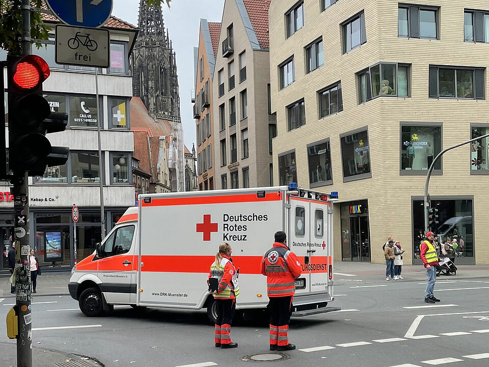 Giro 2025 Zwei Einsatzkräfte des DRK stehen neben einem Krankenwagen an einer Kreuzung, moderne Gebäude und ein Kirchturm im Hintergrund.