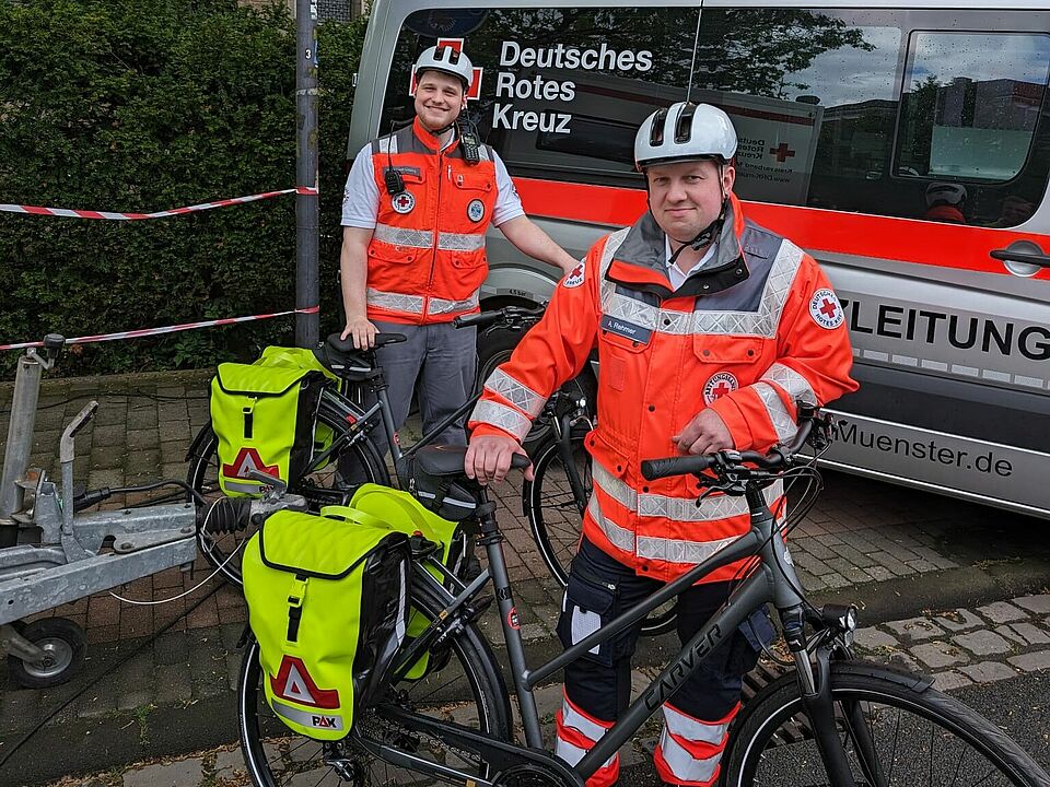 Zwei Einsatzkräfte mit Fahrrädern mit Notfalltaschen. Im Hintergrund ein Einsatzleitfahrzeug.