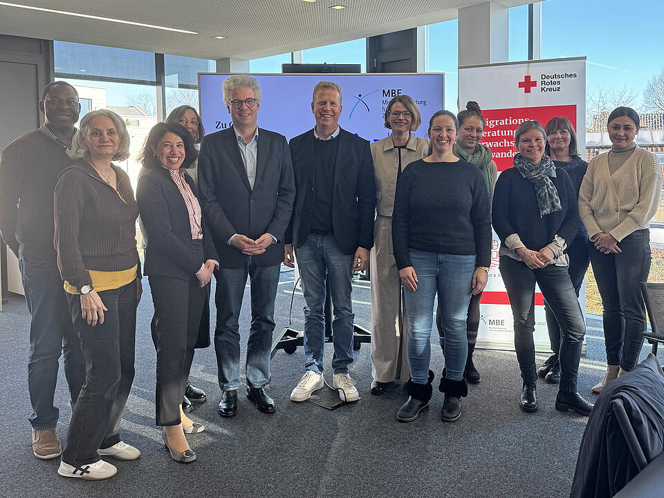 Eine Gruppe von zehn Menschen, die in einem modernen Raum zusammenstehen und lächeln. Im Hintergrund ist ein Banner des Deutschen Roten Kreuzes zu sehen.