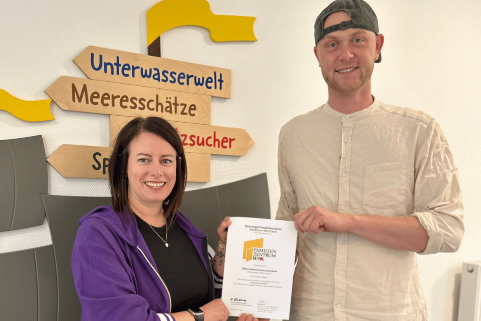 Zwei Personen halten eine Urkunde in die Kamera. Im Hintergrund ein Wegweiser im Schiffsstil zur "Unterwasserwelt" und zu den "Meeresschätzen"