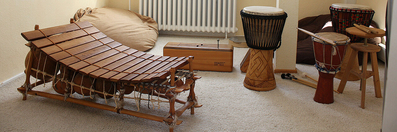 Kinderschutzambulanz Ein Musikraum mit verschiedenen Instrumenten. Im Vordergrund steht ein großes, xylophonähnliches Instrument aus Holz. Dahinter befinden sich mehrere Trommeln, darunter Djemben, sowie ein hölzernes Kasteninstrument. Auf dem Boden liegen Drumsticks und Schlägel. Der Raum hat beige Wände, einen Heizkörper und ist mit Teppichboden ausgelegt