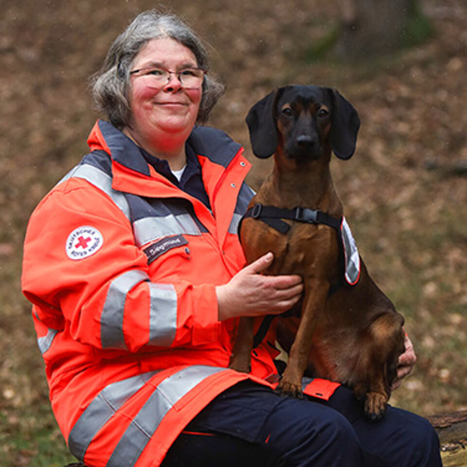 Eine Person in Einsatzkleidung hält einen braunen Hund im Arm, der ein Geschirr trägt. Auf dem Ärmel der Kleidung ist das Logo des Deutschen Roten Kreuzes zu sehen.