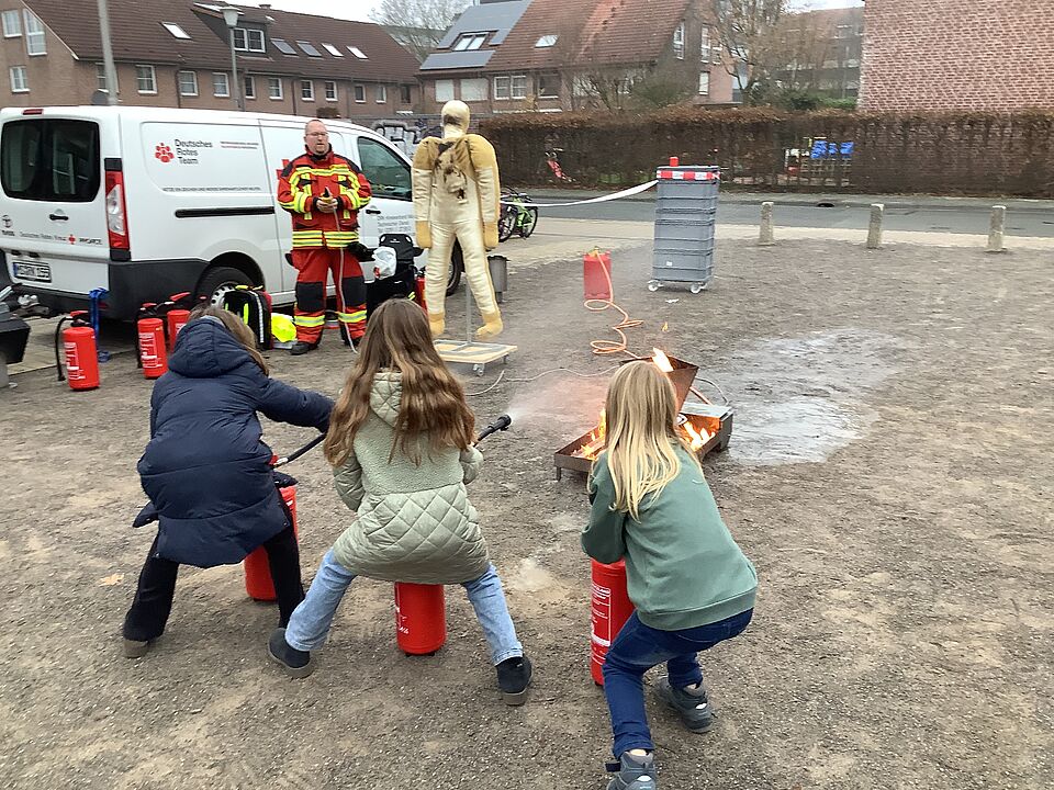 Drei Kinder löschen mit Feuerlöschern ein Feuer, im Hintergrund befindet sich eine Person in Einsatzkleidung und eine Puppe, ein Auto und Anhänger mit DRK-Beschriftung und weitere Feuerlöscher.