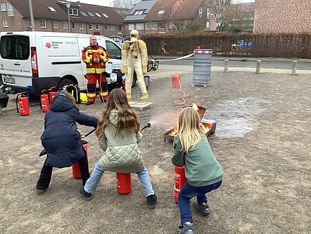 Drei Kinder löschen mit Feuerlöschern ein Feuer, im Hintergrund befindet sich eine Person in Einsatzkleidung und eine Puppe, ein Auto und Anhänger mit DRK-Beschriftung und weitere Feuerlöscher.