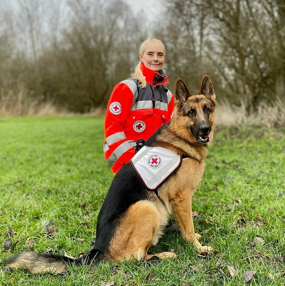 Eine Person in Einsatzkleidung kniet auf einer Wiese. Vor ihr sitzt ein Hund, der eine Weste mit dem Rotkreuz-Logo und dem Aufdruck „Rettung“ trägt. Im Hintergrund sind Bäume zu sehen.