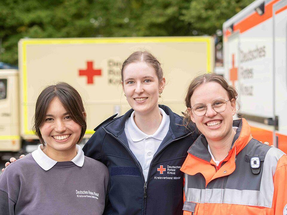 Gruppenfoto von drei Frauen in Einsatzkleidung, im Hintergrund sind DRK-Fahrzeuge zu sehen