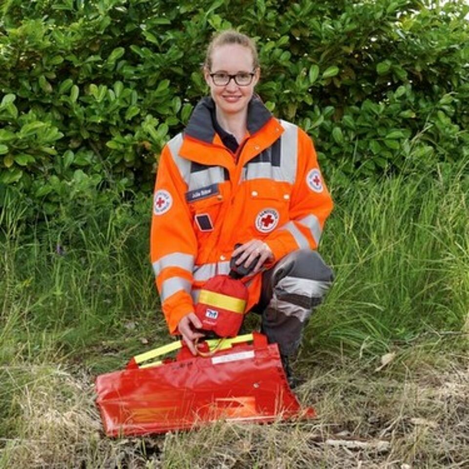 Eine Person in Einsatzkleidung kniet neben einem Notfallrucksack. Im Hintergrund befindet sich grüne Vegetation.