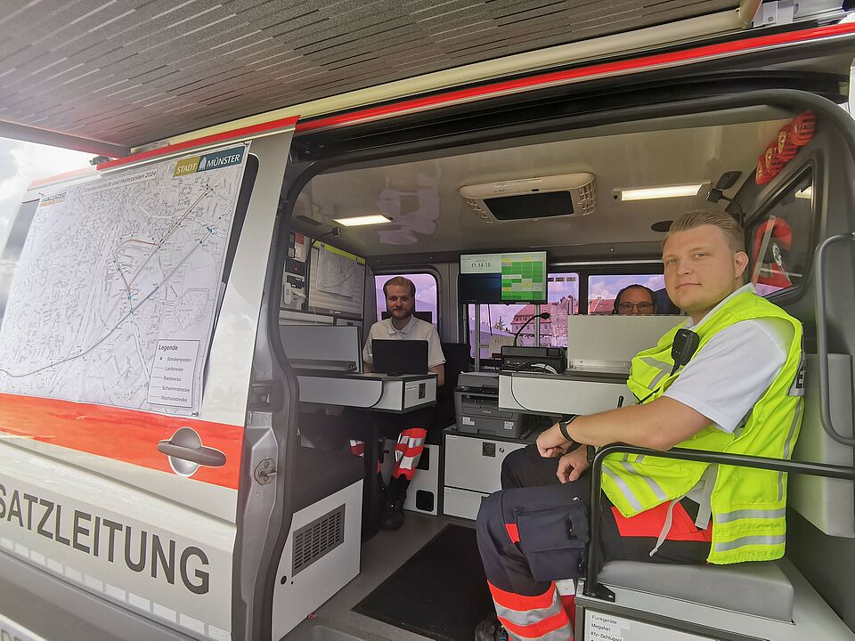 Drei Personen sitzen im Einsatzleitungsfahrzeug, im Hintergrund ist viel Technik und Bildschirme zu sehen.