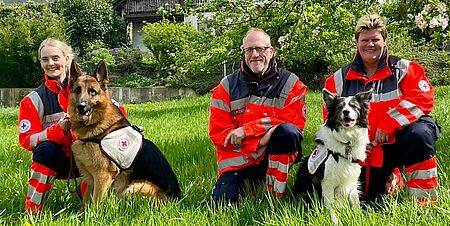 Drei Personen in Einsatzkleidung knien mit zwei Hunden auf einer Wiese. Im Hintergrund ein Baum und Häuser.