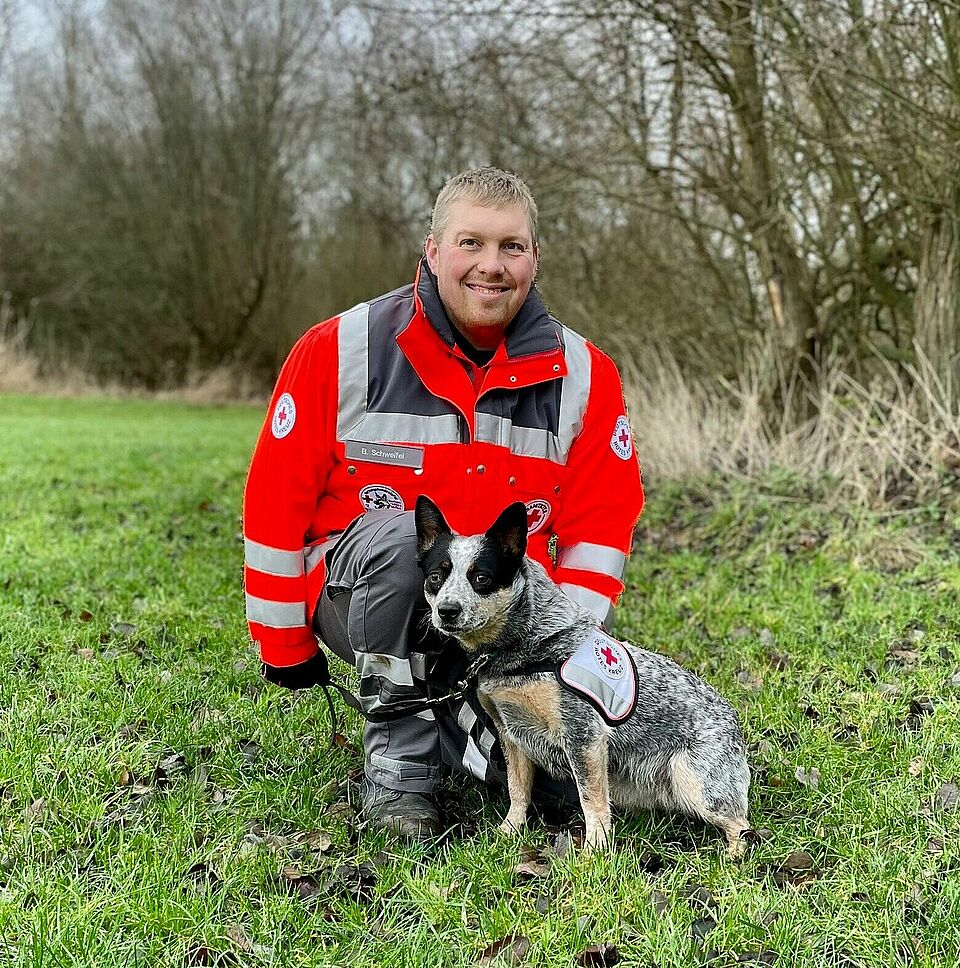 Eine Person in Einsatzkleidung kniet auf einer Wiese neben einem Hund. Der Hund trägt eine Weste mit DRK-Logo. Im Hintergrund sind Bäume und Gras zu sehen