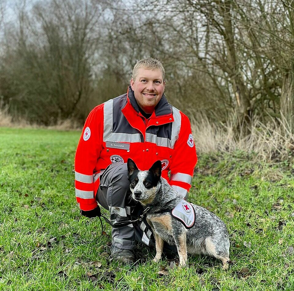 Eine Person in Einsatzkleidung kniet auf einer Wiese neben einem Hund. Der Hund trägt eine Weste mit DRK-Logo. Im Hintergrund sind Bäume und Gras zu sehen
