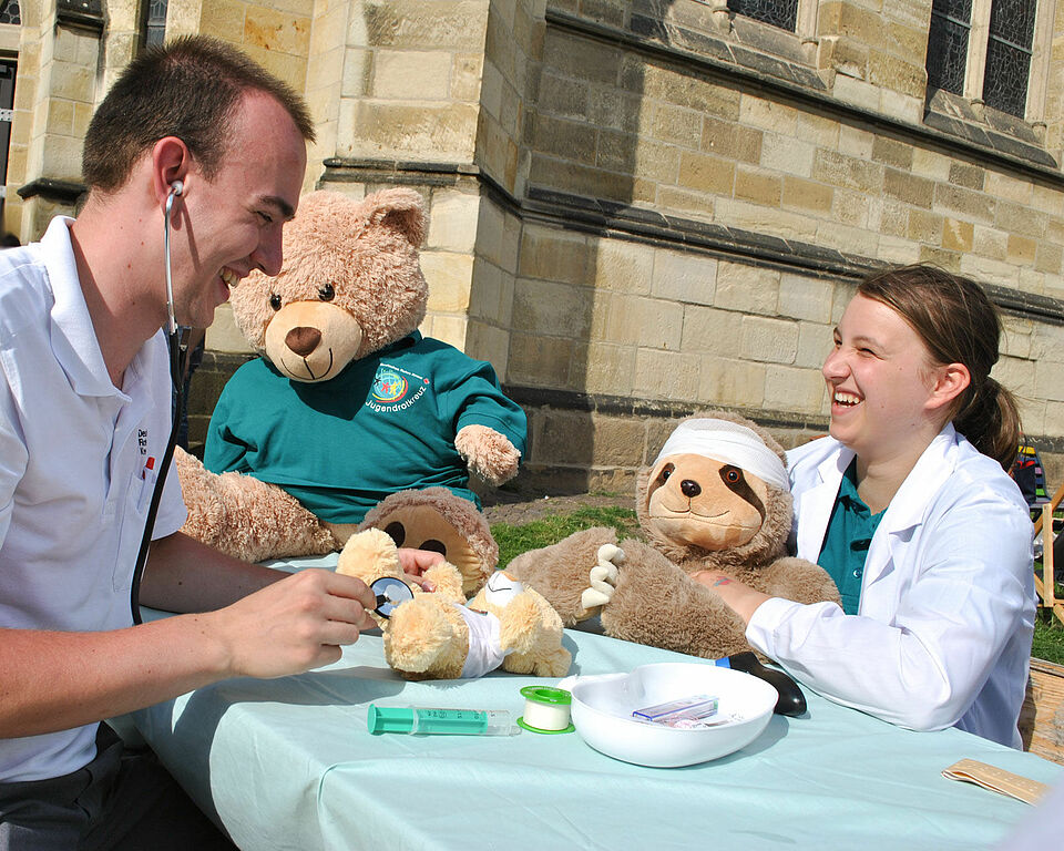 Zwei Personen sitzen an einem Tisch, auf dem Teddys der Teddyklinik liegen. Eine Person hört mit einem Stetoskop einen Teddy ab, die andere Person lacht