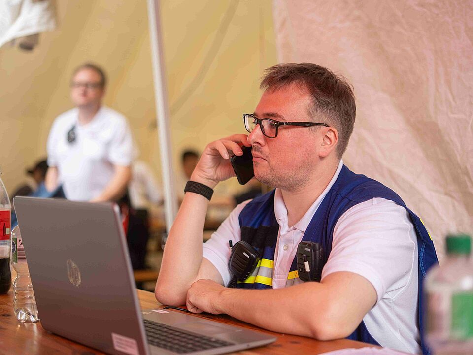 Eine Person mit blauer Weste sitzt an einem Tisch vor einem Laptop und telefoniert.