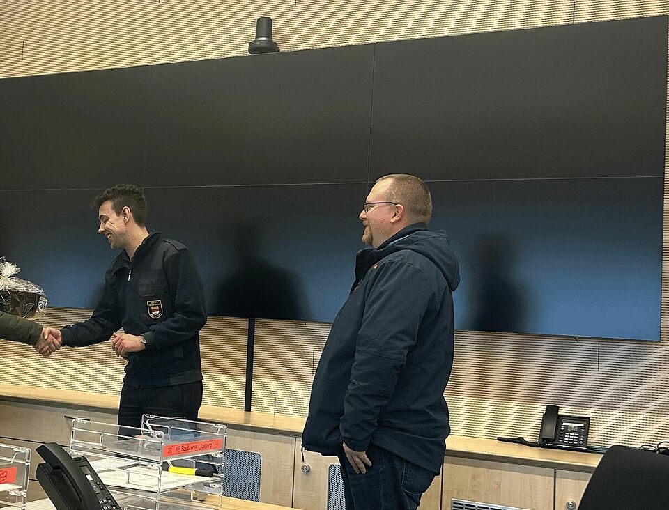 Drei Personen stehen vor einem großen Bildschirm. Eine Person übergibt einer anderen Person ein Geschenk und schüttelt ihr die Hand.