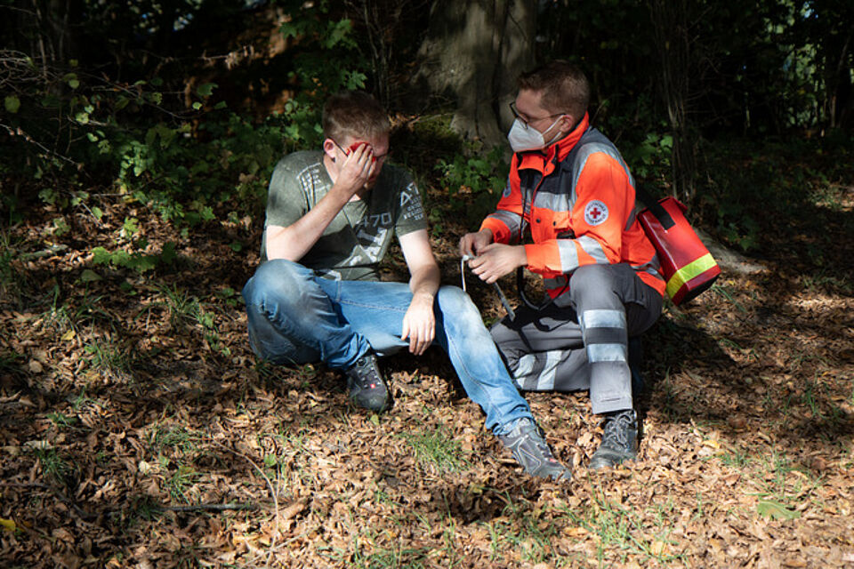Zwei Personen sitzen auf dem Boden in einem bewaldeten Gebiet. Eine Person trägt Freizeitkleidung mit grünem T-Shirt und blauer Jeans und hält sich eine Hand vors Gesicht. Die andere Person trägt Einsatzkleidung. Diese Person scheint der anderen Erste Hilfe zu leisten.