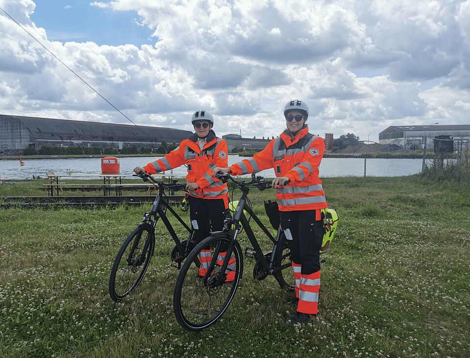Zwei Personen in Einsatzkleidung mit Fahrrädern auf einer Wiese, im Hintergrund ein Kanal und Industrie